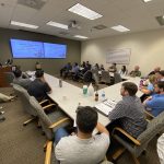 Man in blue shirt giving a speech to a group of people in a conference room